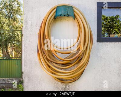 Long weathered garden hose hanging on a white wall during daytime Stock Photo