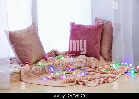 Beautiful glowing garland with pillows and plaid on window sill Stock Photo