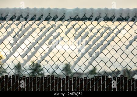 A selective focus view of a chain link diamond mesh fence creating a barrier during winter, covered in white snow and ice against a muted sky Stock Photo