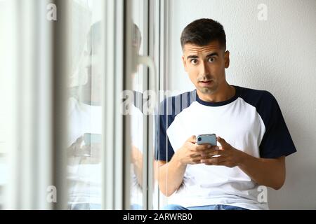 Surprised young man with mobile phone near window Stock Photo