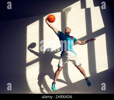 Young athletic basketball player jumping with ball in front of wall with shadows from window Stock Photo