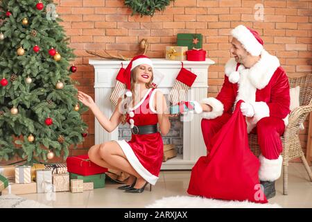 Young couple dressed as Santa Claus putting gifts under fir tree in room decorated for Christmas Stock Photo