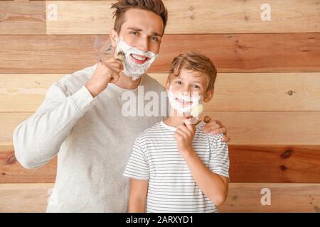 Father with son applying shaving foam onto their faces against wooden background Stock Photo