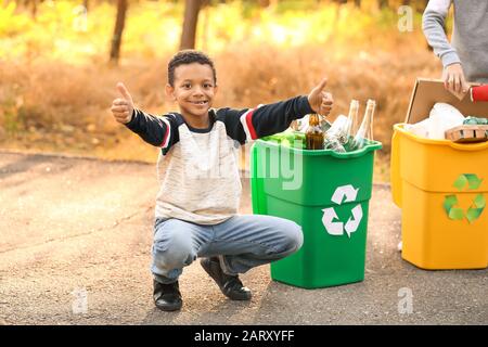 Little African-American boy showing thumb-up near containers for trash outdoors. Concept of recycling Stock Photo