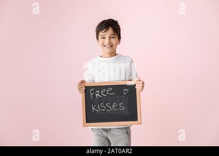 Cute little boy holding chalkboard with written text FREE KISSES on color background. Valentines Day celebration Stock Photo