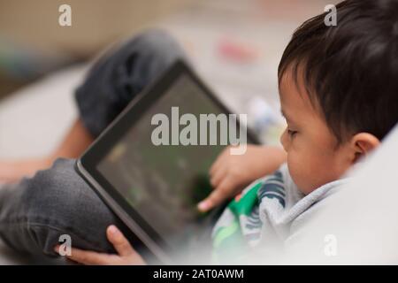 Unsupervised toddler using a tablet at home who is relaxed sitting on a couch and using his finger on the touchscreen. Stock Photo