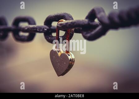 A romantic closed and rusty heart shaped padlock hanging from a chain beside a river with blurred cityscape in the background. Concept of long lasting Stock Photo