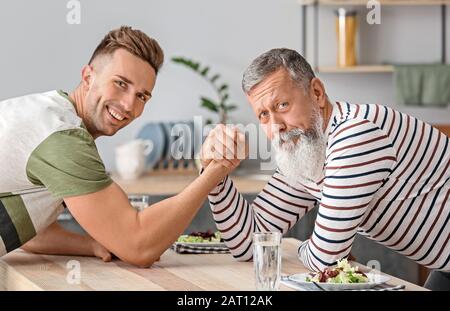 Senior man and his adult son having arm wrestling competition in kitchen Stock Photo