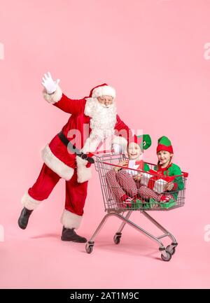 Santa Claus and little elf kids with gifts sitting in shopping cart on color background Stock Photo