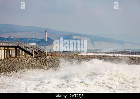 storm waves on the background of the lighthouse and high cliffs. In the foreground an abandoned shingle beach, a promenade with balustrade, stairs. In Stock Photo