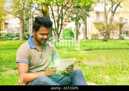 Young indian man having video call on tablet in park university camp - Concept of new technology miracle addiction - Soft warm saturated filter Stock Photo