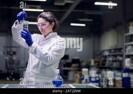 Brunette lab girl in glasses and white coat with experimental glass in her hands conducts experiments on defocused background Stock Photo