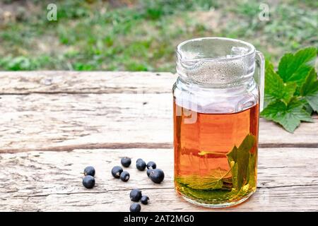 Tea in a glass teapot with green leaves of black currant on a wooden table against the background of green grass. Close-up. Stock Photo