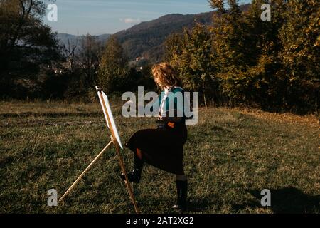 oung female artist working on painting outdoors, in the autumn landscape. She is in front of the canvas and drawing.She holds oil paints, artist brush Stock Photo