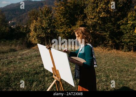 oung female artist working on painting outdoors, in the autumn landscape. She is in front of the canvas and drawing.She holds oil paints, artist brush Stock Photo