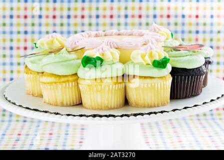 Front view of a birthday cake and cupcakes in pastel colored butter cream icing on a cake stand. The background is a checkered pattern tablecloth. Stock Photo