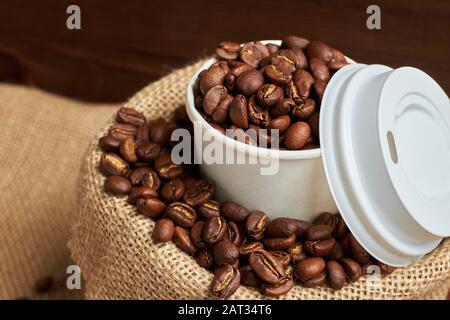 White cardboard cup with coffee beans in a knitted bag on a burnt wooden background. Close up. Stock Photo