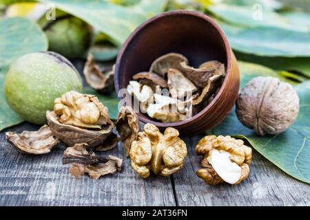 Walnut kernels lie in a bowl next to nuts in green shells and green leaves on a rustic old wooden table. Harvest walnuts. Stock Photo