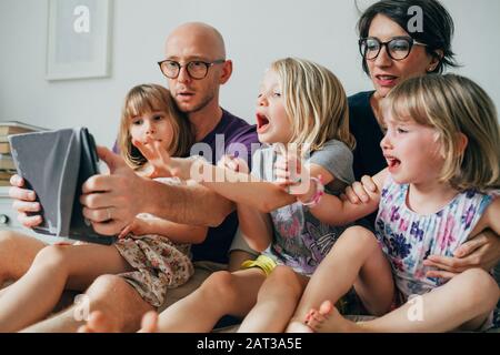Three little sisters indoor at home using tablet lying in bed supervised by parents - education, security, streaming concept Stock Photo