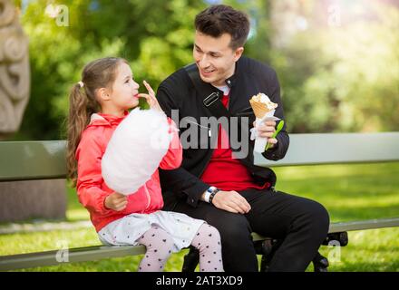 Cute girl and father eating cotton candy and ice cream, sitting on bench in park. Happy family having quality time together Stock Photo