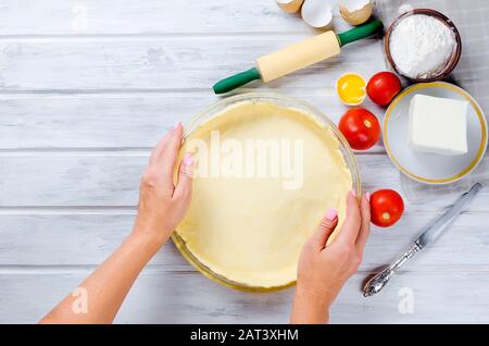 Raw Shortbread dough for baking tart with tomatoes and cheeses filling in glass round baking form on white wooden table and womans hands holding bakin