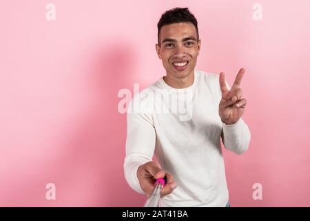 Happy mulatto man taking a self portrait with selfie stick ahnd showing a victoty gesture against a pink background. - Image Stock Photo