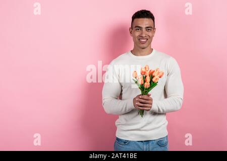 Happy mulatto man standing against a pink background with tulips bouquet in his hands. - Image Stock Photo
