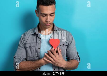 Mulatto man sadly looking at the heart-object holding in his hands it and leaning it against his chest. Blue studio background - Image Stock Photo