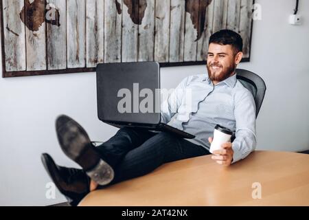 Nice guy with a beard sitting at a table, putting his feet on the table, drinking coffee and working on a computer Stock Photo