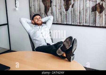 An attractive guy with a beard puts his hands behind his head, puts his feet on the table and rests. Stock Photo