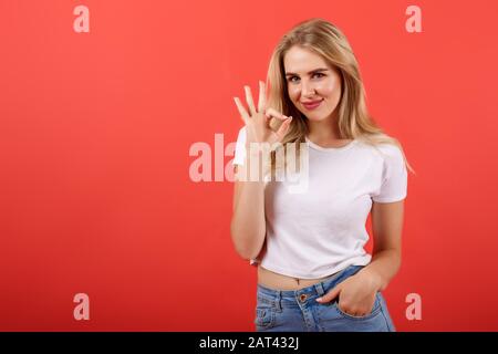 Cheerful pretty girl young cute pretty woman with blonde hair in t-shirt shows ok gesture on red background Stock Photo