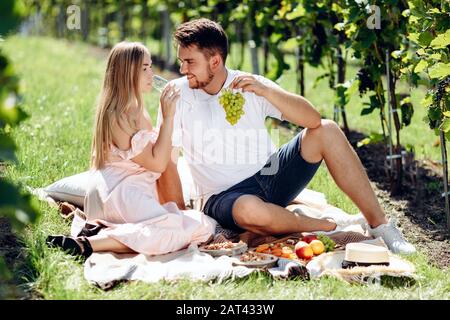 Enamored girl and boy sitting on blanket eating grapes and drinking wine during picnic in grape garden Stock Photo