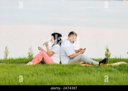 Senior woman and  senior man sitting on a grass. Woman drinking a water and man in earphones looking to phone.- Image Stock Photo