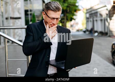 Senior businessman holding open laptop and attentively looking at the screen touches his face- Image Stock Photo