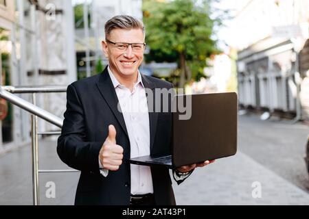 Happy businessman holding open laptop, smiling with thumb up- Image Stock Photo