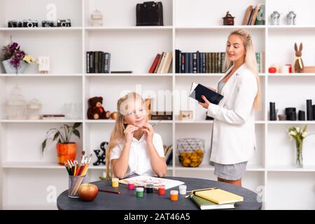 Dreamy, smiling girl draws at the table, her mother watching in the background. Stock Photo