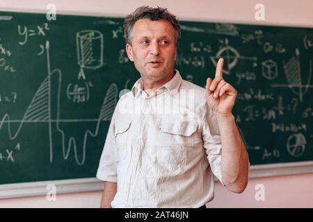 Teacher stands next the blackboard and explain a lesson holding his index finger up.- Image Stock Photo