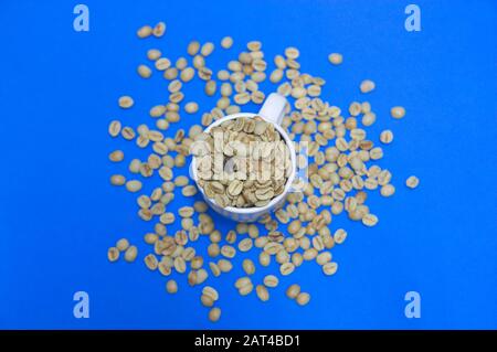 A cup of coffee full of raw, unroasted coffee beans seen from above. In addition, coffee beans are also scattered on the table that is classic blue Stock Photo