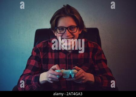 Happy teenage boy playing video games seated in his armchair. Excited adolescent wearing eyeglasses, holding joystick console smiling to camera, try t