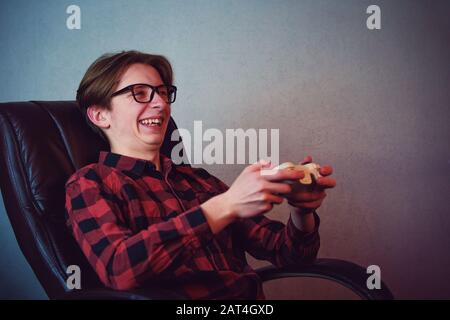 Happy teenage boy playing video games seated in his armchair. Excited adolescent wearing eyeglasses, holding joystick console smiling to camera, try t