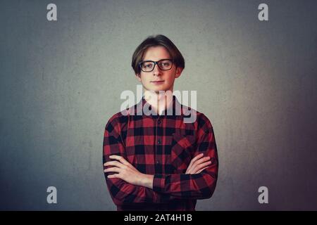 Positive teenage guy hipster, wearing casual red shirt and glasses, keeps hands folded looking serious to camera isolated on grey wall background. Stock Photo