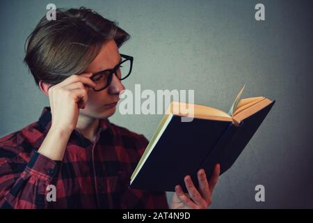 Close up portrait surprised student boy, fixing glasses, double check a information from the book. Shocked reader being confused isolated on grey wall Stock Photo