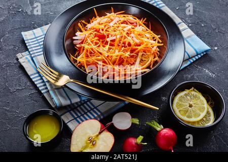 close-up of Fresh carrot radish and apple salad sprinkled with quinoa seeds and lemon zest in a black bowl on a wooden table with homemade dressing Stock Photo