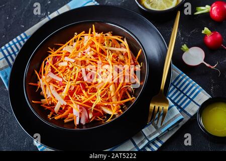 close-up of Fresh carrot radish and apple salad sprinkled with quinoa seeds and lemon zest in a black bowl on a wooden table with homemade dressing Stock Photo