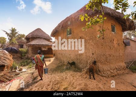 Rural Indian village scene with woman carrying bucket of water with view of mud huts and unpaved village road near Bolpur West Bengal Stock Photo