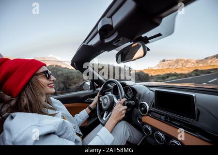 Happy woman in red hat driving convertible car while traveling on the desert road. Carefree lifestyle and travel concept