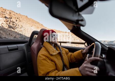 Happy woman in bright hat and jacket driving convertible car while traveling on the desert road on a sunset. Image focused on the background, woman is out of focus Stock Photo