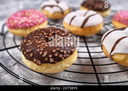 Tasty baked donut with brown chocolate glazing and sprinkles on cake grid with other colorful donuts in background Stock Photo