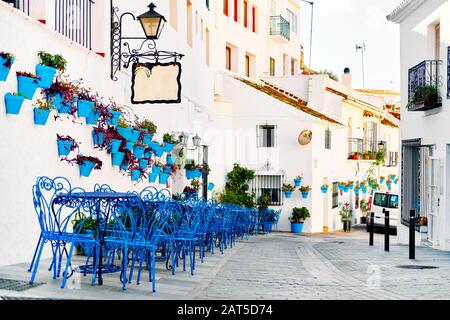 Mijas Pueblo Blanco, charming small village, picturesque empty street in old town with bright blue tables chairs of local cafe, flower pots hanging on Stock Photo
