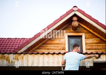 a man in a white T-shirt performs repairs on the roof of a wooden house. View from the back Stock Photo